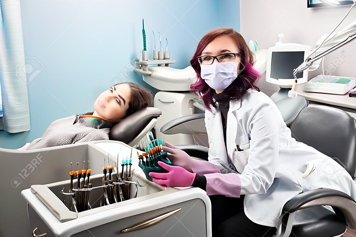 Női fogorvos nő beteg a székre a fogorvosi rendelőben. Orvos visel szemüveget, maszkot, fehér egyenruhát és rózsaszín kesztyűt. Fogászat. Fogászati felszerelések