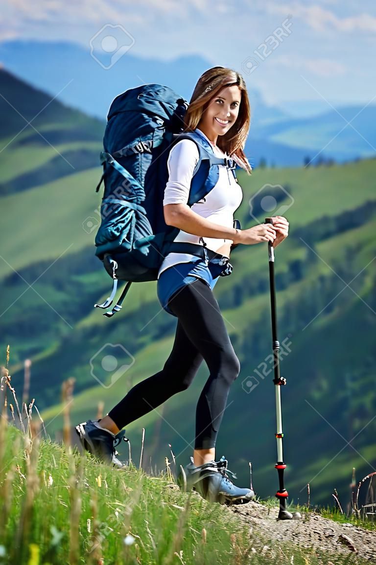 Full-length portrait of beautiful sporty girl hiker with blue backpack and trekking poles posing on the top of a hill, smiling to the camera in the mountains. Concept of active lifestyle