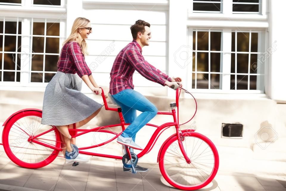 Young romantic pair, handsome man and pretty blond woman cycling together tandem double red bike along paved sidewalk on bright sunny summer day by old buildings