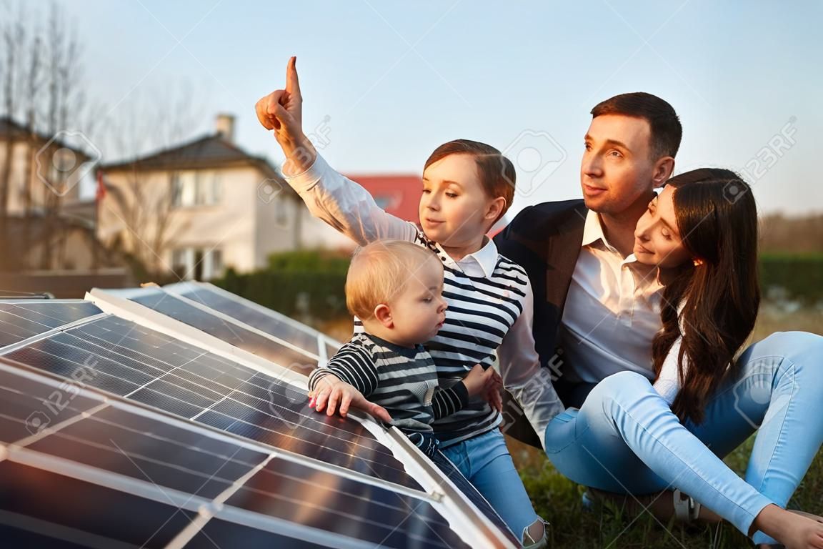 El hombre le muestra a su familia los paneles solares en la parcela cerca de la casa durante un día caluroso. Mujer joven con un niño y un hombre en los rayos del sol mirar los paneles solares.