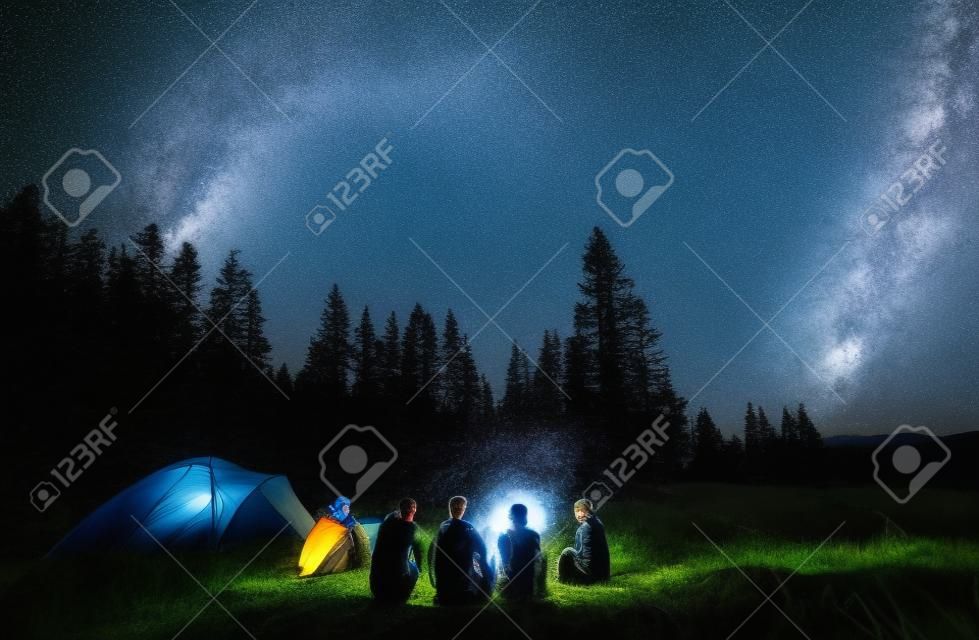 Noite de verão acampando nas montanhas, floresta de abeto no fundo, céu com estrelas e maneira leitosa. Grupo de vista traseira de cinco turistas que têm um descanso juntos em torno da fogueira, desfrutando de ar fresco perto da barraca.