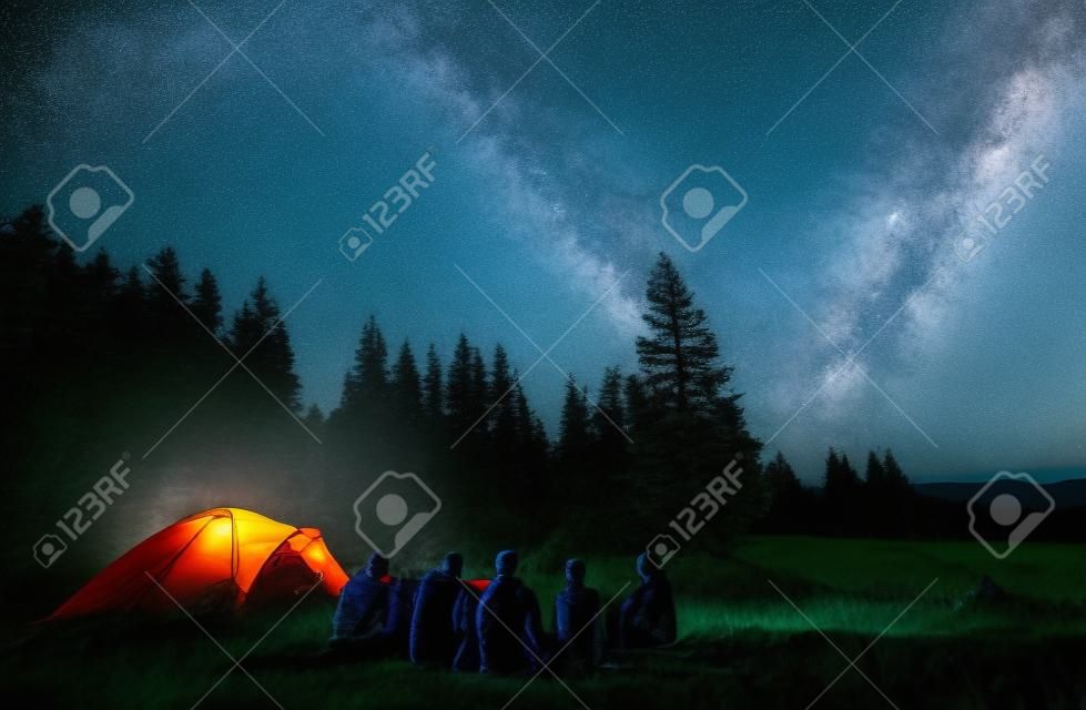 Noite de verão acampando nas montanhas, floresta de abeto no fundo, céu com estrelas e maneira leitosa. Grupo de vista traseira de cinco turistas que têm um descanso juntos em torno da fogueira, desfrutando de ar fresco perto da barraca.