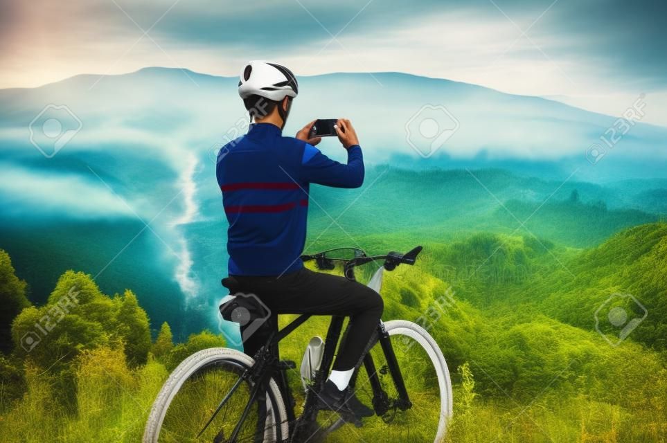 Rückansicht eines Mannes, der auf dem Fahrrad sitzt und ein Bergfoto mit dem Smartphone spricht. Männlicher Radfahrer, der Handy hält und Foto von schönen nebligen Hügeln macht. Konzept von Sport, Radfahren und Fotografie.