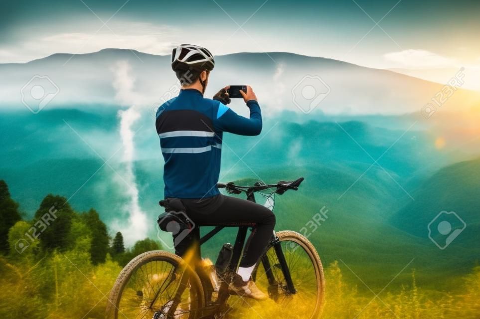 Rückansicht eines Mannes, der auf dem Fahrrad sitzt und ein Bergfoto mit dem Smartphone spricht. Männlicher Radfahrer, der Handy hält und Foto von schönen nebligen Hügeln macht. Konzept von Sport, Radfahren und Fotografie.
