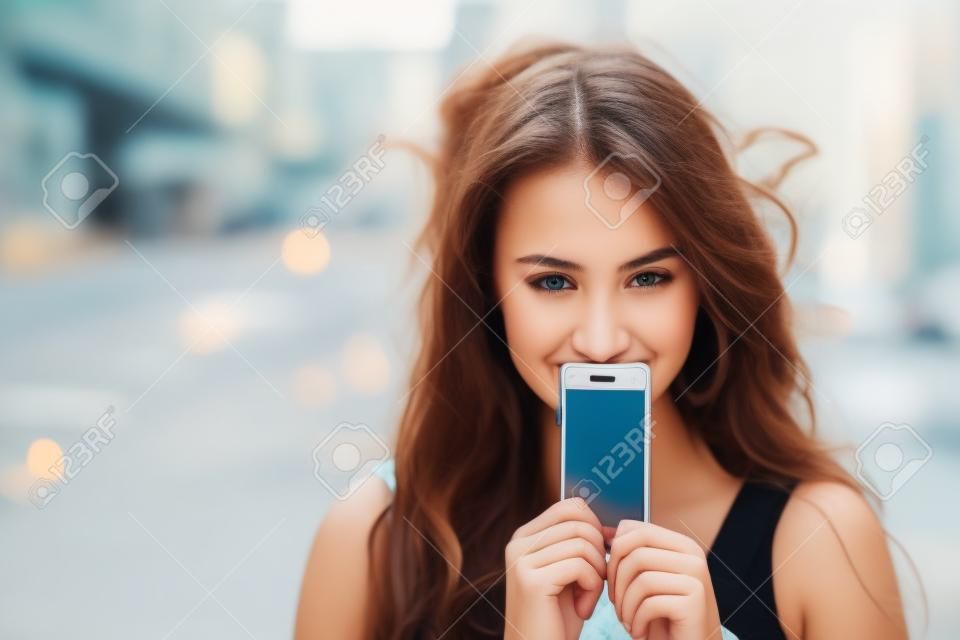 一个年轻女人在一个背景的夏天街道上覆盖她的屏幕智能手机