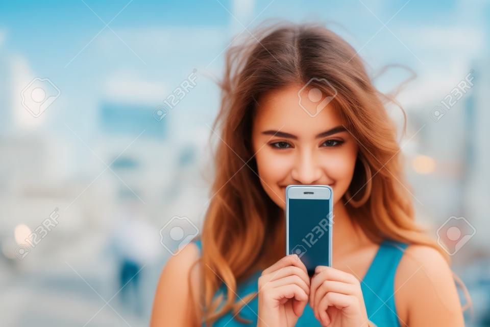 一个年轻女人在一个背景的夏天街道上覆盖她的屏幕智能手机