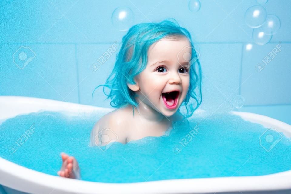 Divertente allegro bambino felice che fa il bagno giocando con le bolle di schiuma. Piccolo bambino in una vasca da bagno. Bambino sorridente in bagno su sfondo blu. Igiene e assistenza sanitaria