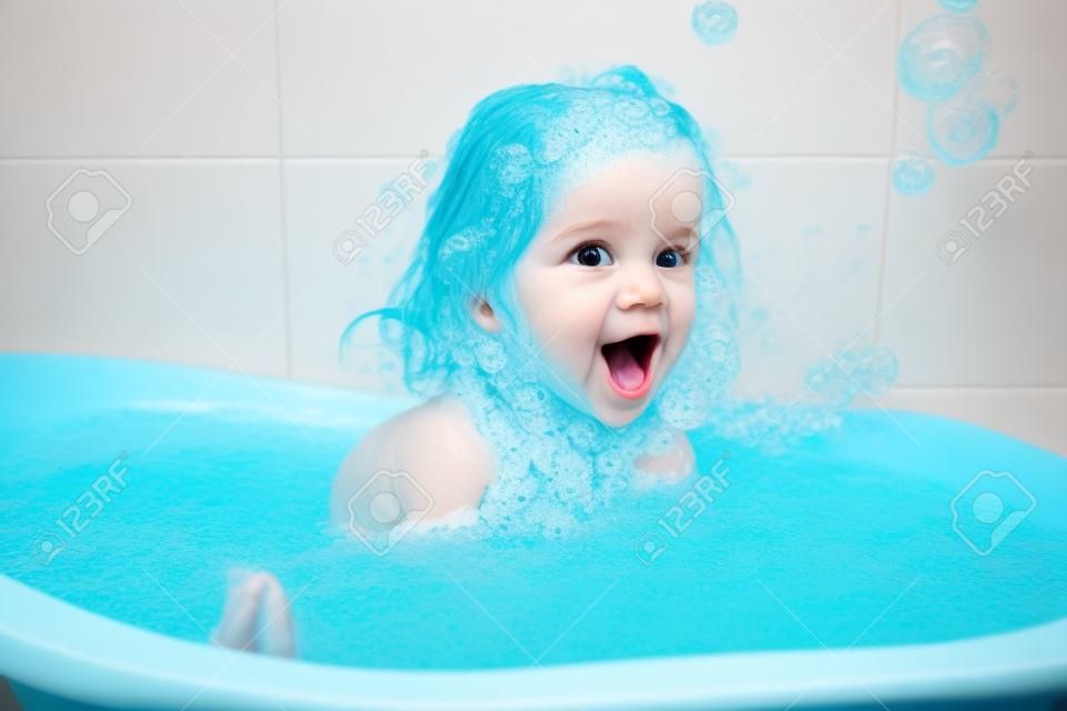 거품 거품을 가지고 노는 목욕을하는 재미 쾌활한 행복한 유아 아기. 욕조에 있는 어린 아이. 파란색 배경에 욕실에서 웃는 아이. 위생 및 건강 관리