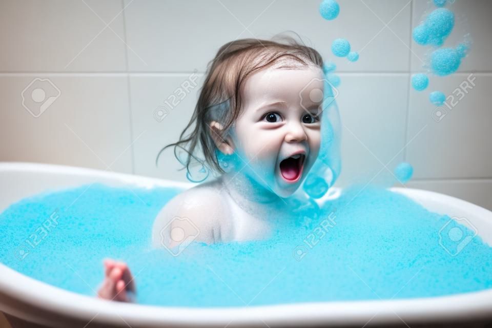 Spaß fröhliches glückliches Kleinkindbaby, das ein Bad nimmt und mit Schaumblasen spielt. Kleines Kind in einer Badewanne. Lächelndes Kind im Badezimmer auf blauem Hintergrund. Hygiene und Gesundheitsvorsorge