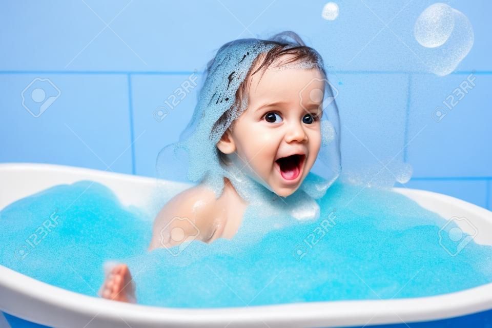 Fun cheerful happy toddler baby taking a bath playing with foam bubbles. Little child in a bathtub. Smiling kid in bathroom on blue background. Hygiene and health care