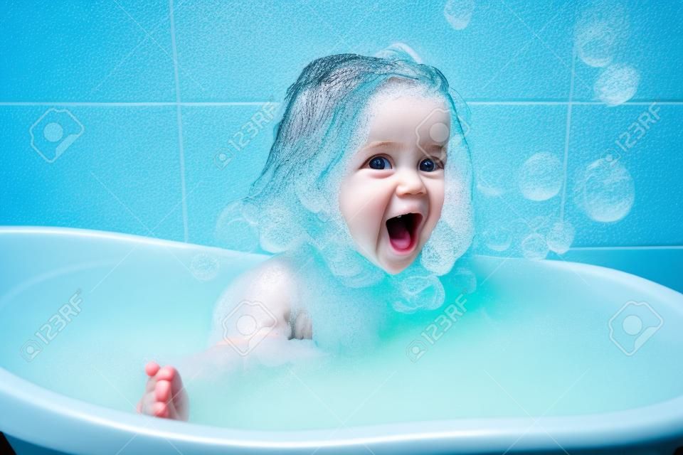 Fun cheerful happy toddler baby taking a bath playing with foam bubbles. Little child in a bathtub. Smiling kid in bathroom on blue background. Hygiene and health care