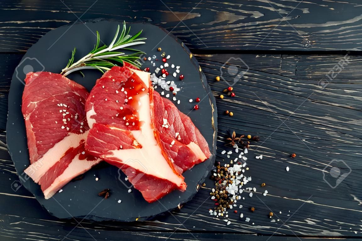 Three pieces of juicy raw beef with rosemary on a stone cutting board on a black wooden table background. Meat for barbecue or grill sprinkled with pepper and salt seasoning