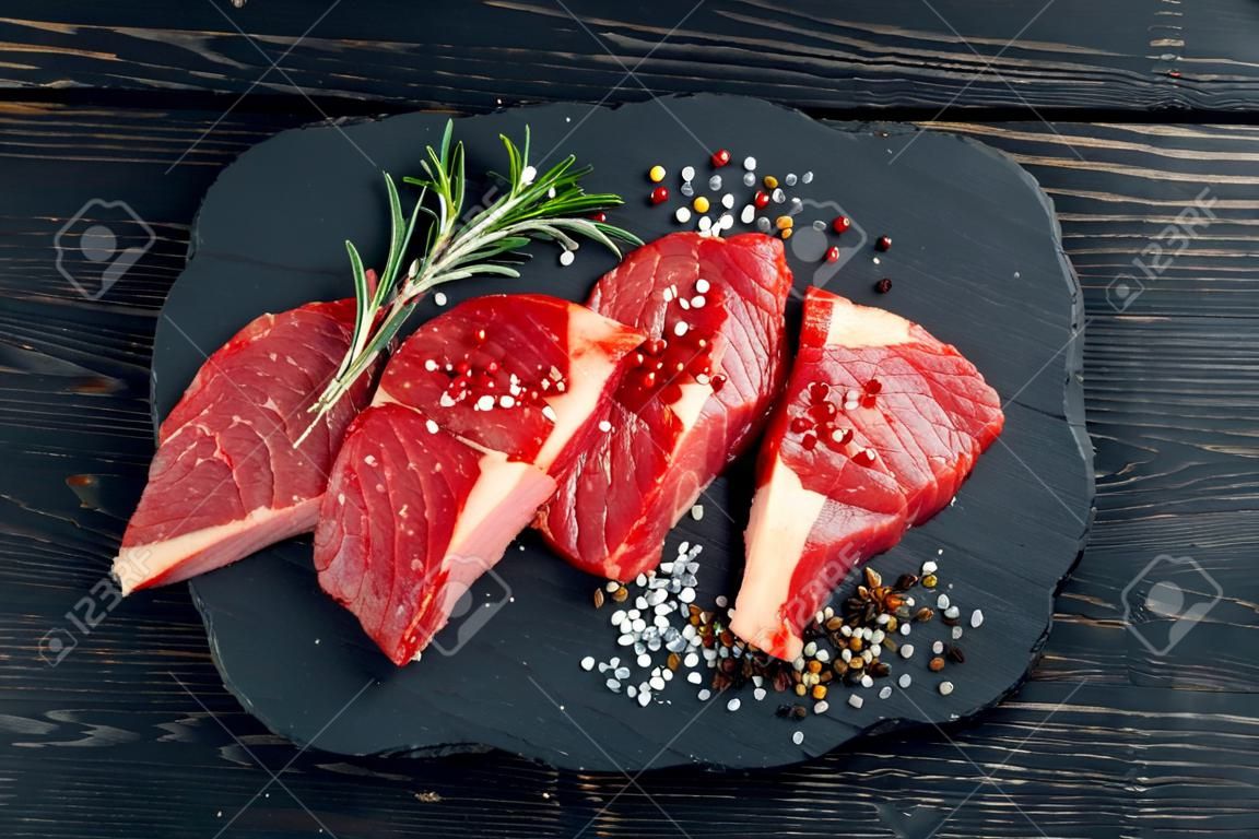 Three pieces of juicy raw beef with rosemary on a stone cutting board on a black wooden table background. Meat for barbecue or grill sprinkled with pepper and salt seasoning
