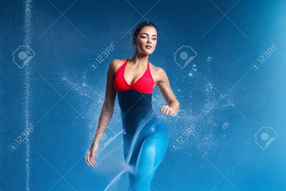Piękna młoda dziewczyna w czerwonej odzieży sportowej w aqua studio. krople wody rozlały się po jej sprawnym ciele. idealna figura na tle rozprysków wody. klasyczne ciemnoniebieski kolor tła