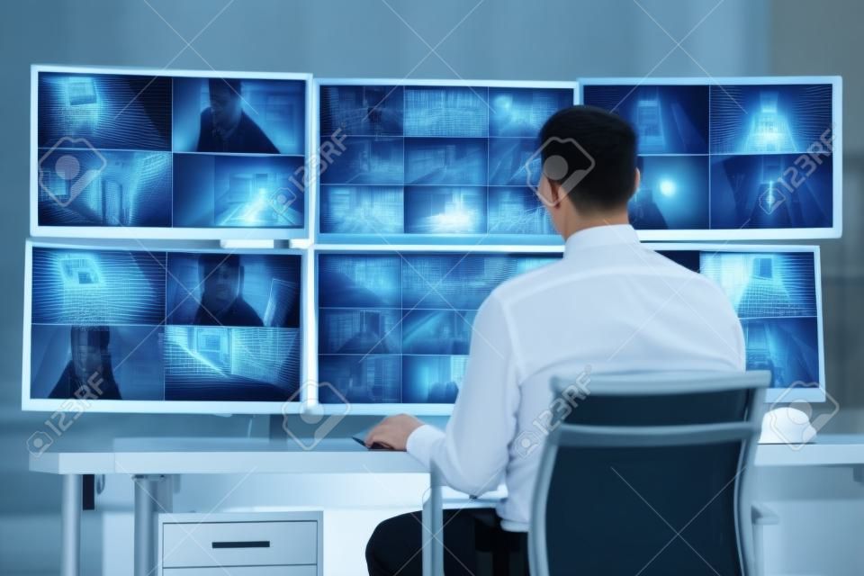 Rear view of security system operator looking at CCTV footage at desk in office