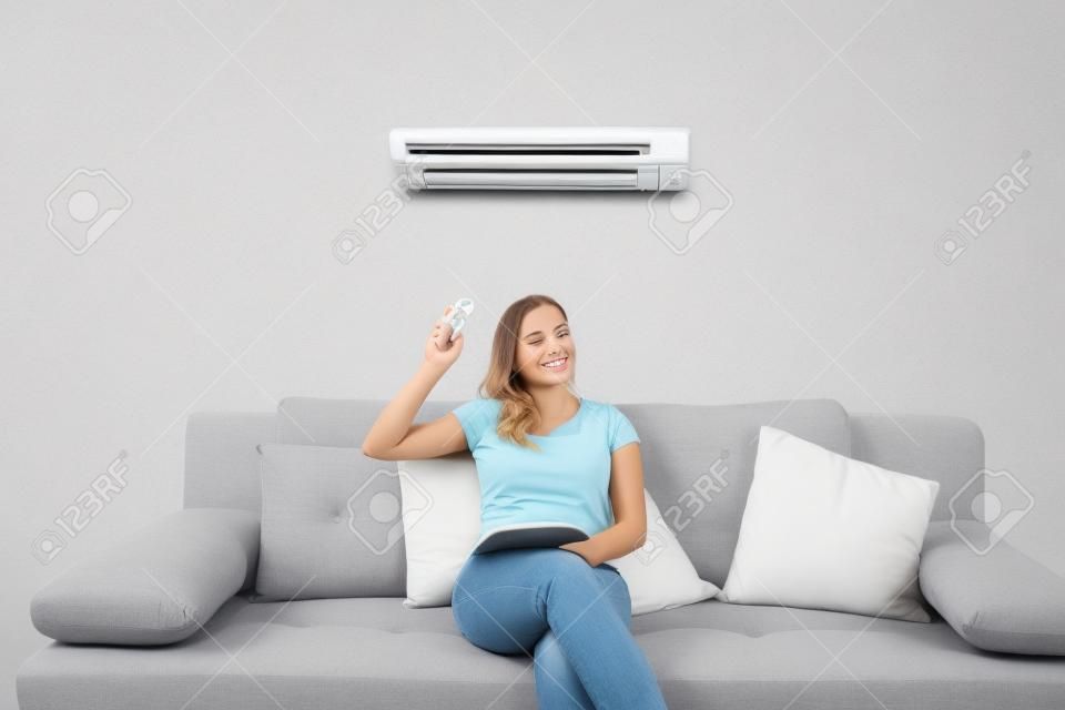 Jeune femme heureuse assise sur le climatiseur fonctionnant avec télécommande à la maison