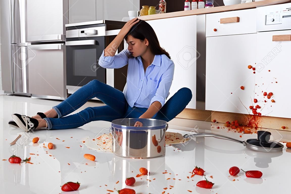 台所でこぼれた食べ物で台所の床に座っている不幸な女性