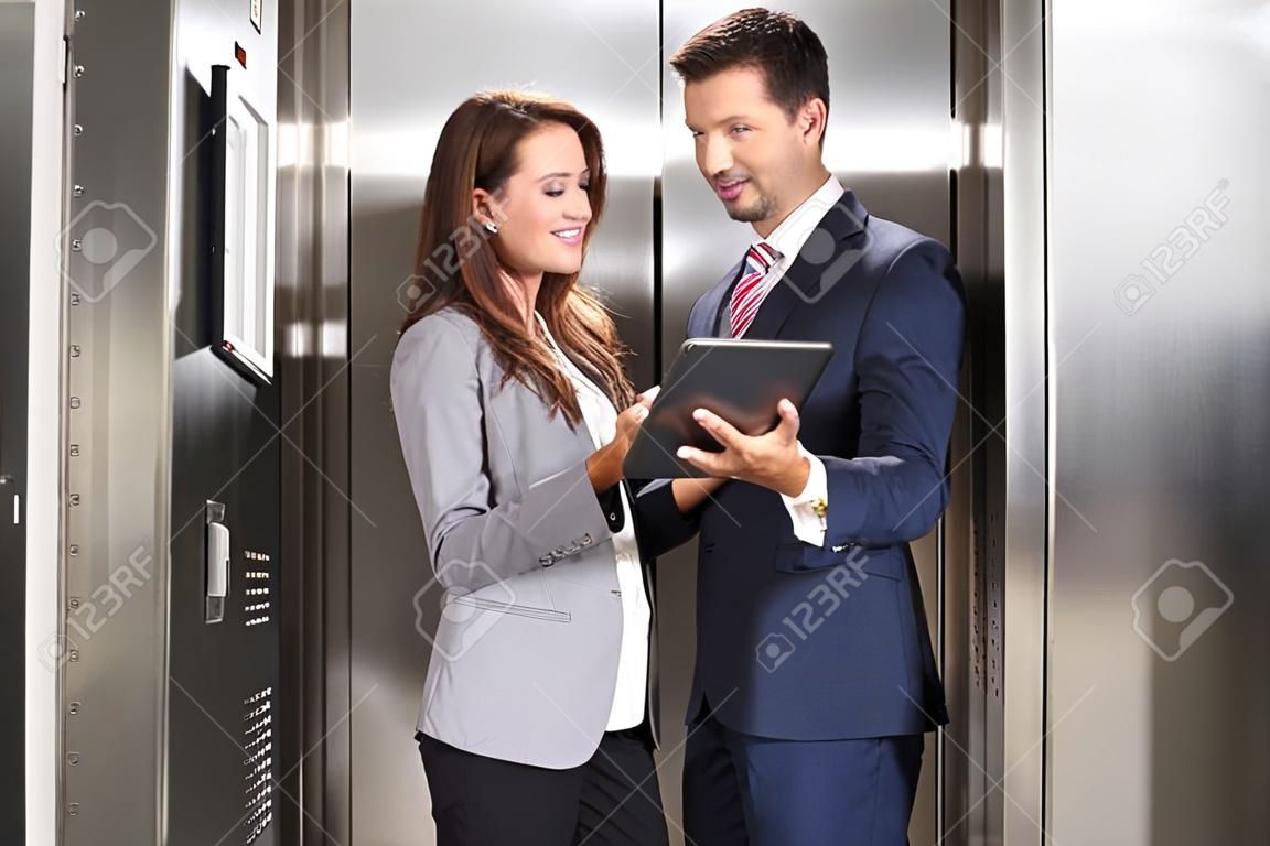 エレベーターの近くに立っているデジタルタブレットを使用しながら議論する笑顔の若いビジネスウーマンとビジネスマン