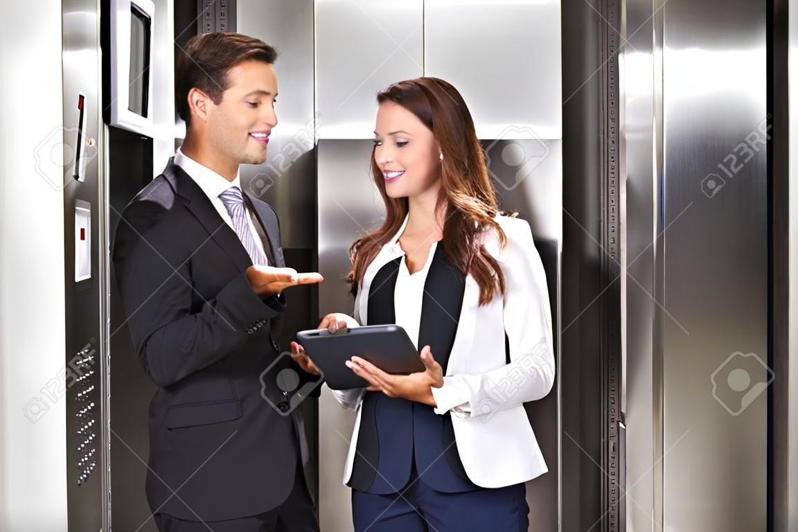 Sonriente joven empresaria y empresario discutiendo mientras usa tableta digital de pie cerca del ascensor