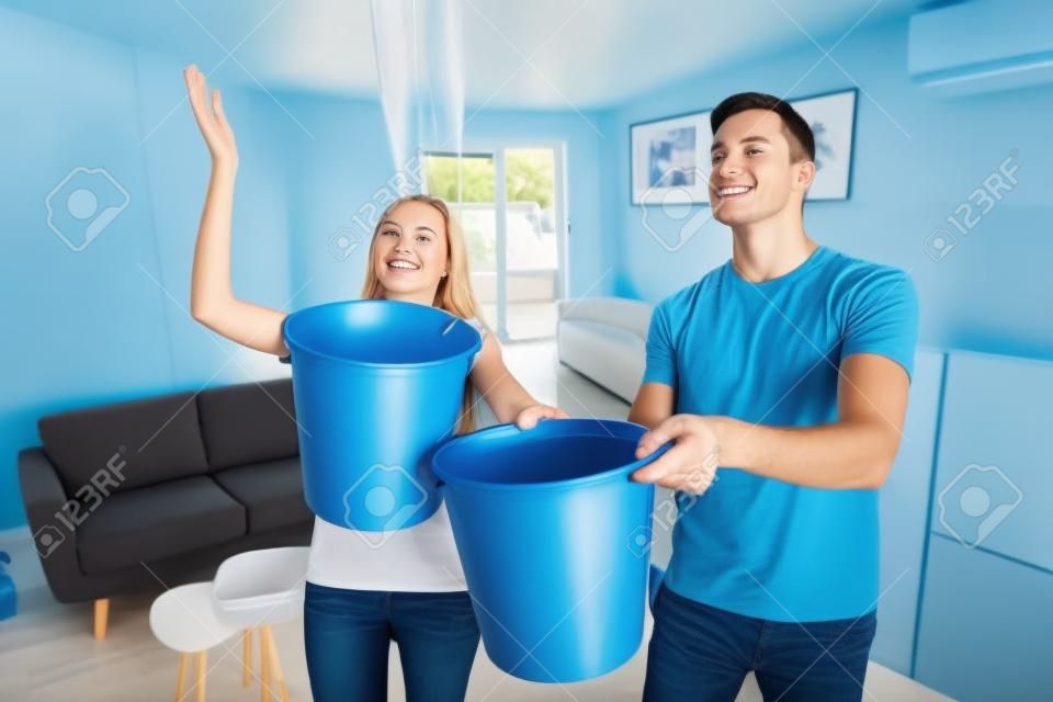 Joven esposo y esposa con balde azul recolectando agua del techo dañado en la sala de estar