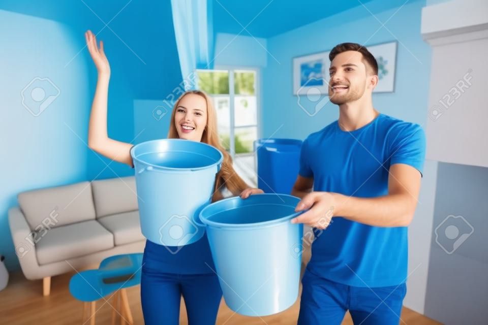 거실의 손상된 천장에서 물을 모으는 파란색 양동이를 가진 젊은 남편과 아내