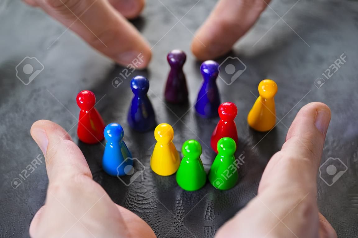 Close-up Of A Person's Hand Protecting Multicolored Pawns Forming Circle Over White Desk