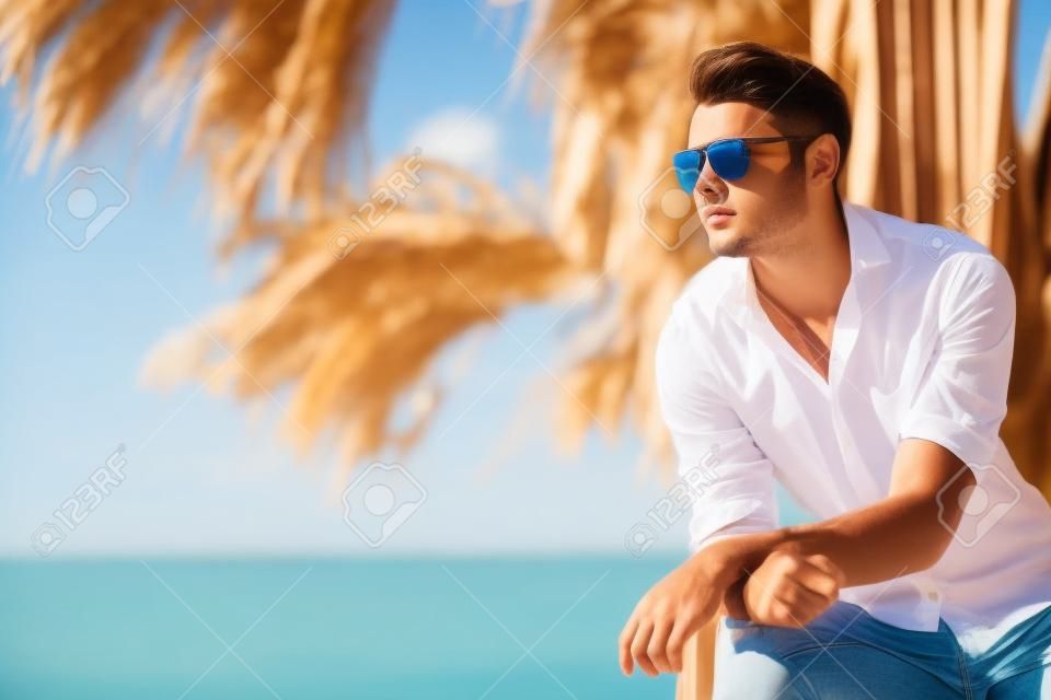 Young attractive man with sunglasses looking out over the sea during the summer. He looking forward, dressed in a white shirt and leaning on a wooden construction.