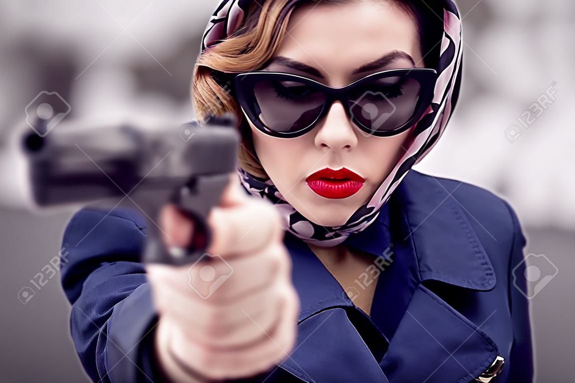 Schöne brünette Spionageagentin (Mörder oder Polizei) in Jacke und Sonnenbrille mit einer Waffe in der Hand.
