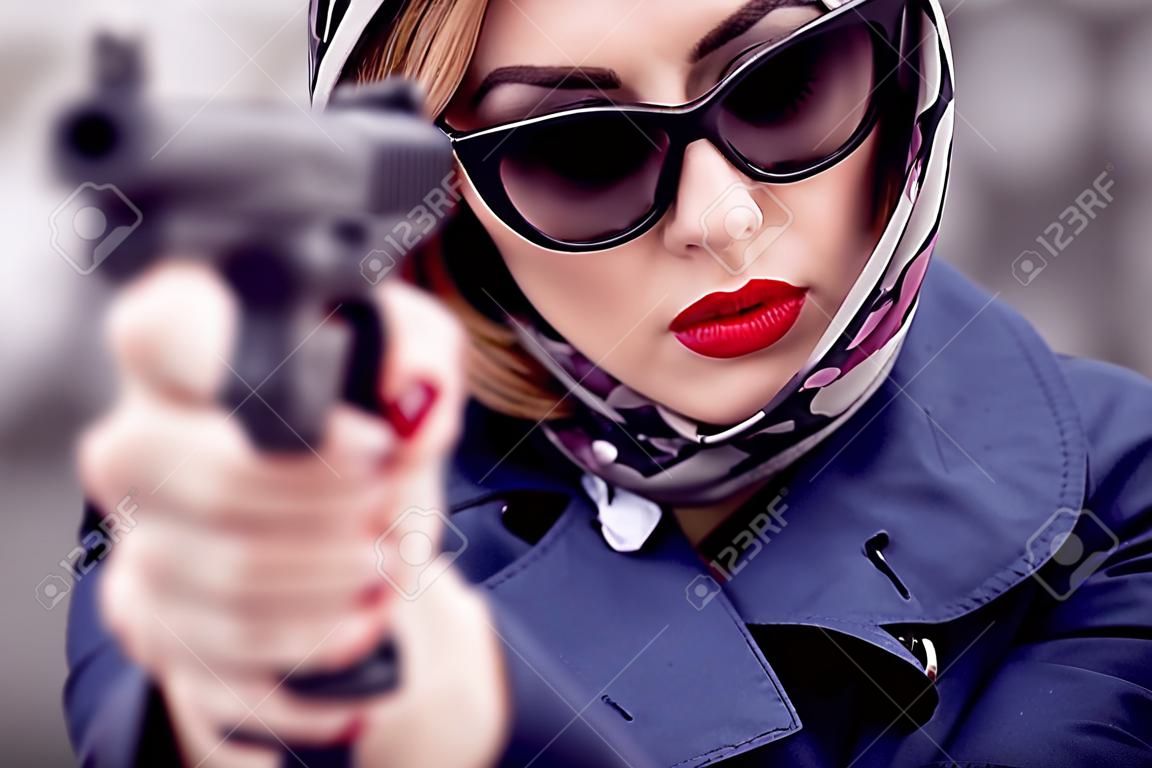 Schöne brünette Spionageagentin (Mörder oder Polizei) in Jacke und Sonnenbrille mit einer Waffe in der Hand.