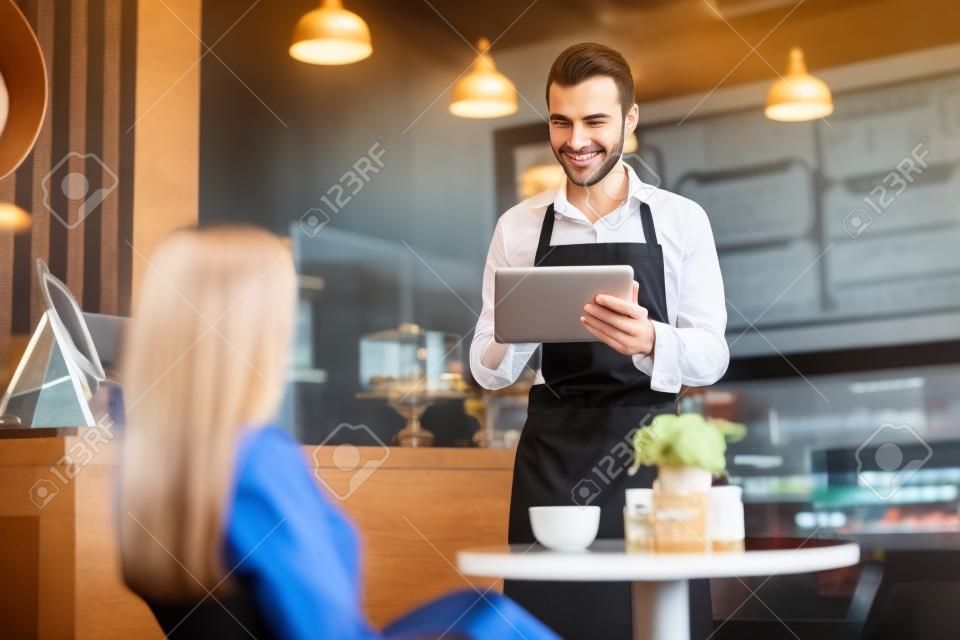 Красивый молодой официант принимает заказ клиента с помощью планшетного компьютера в кафе