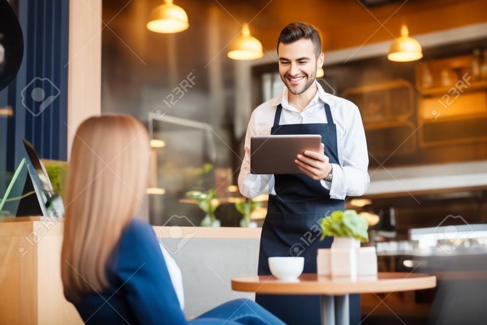 Przystojny młody kelner biorąc zamówienie klienta przy użyciu komputera typu tablet w kawiarni