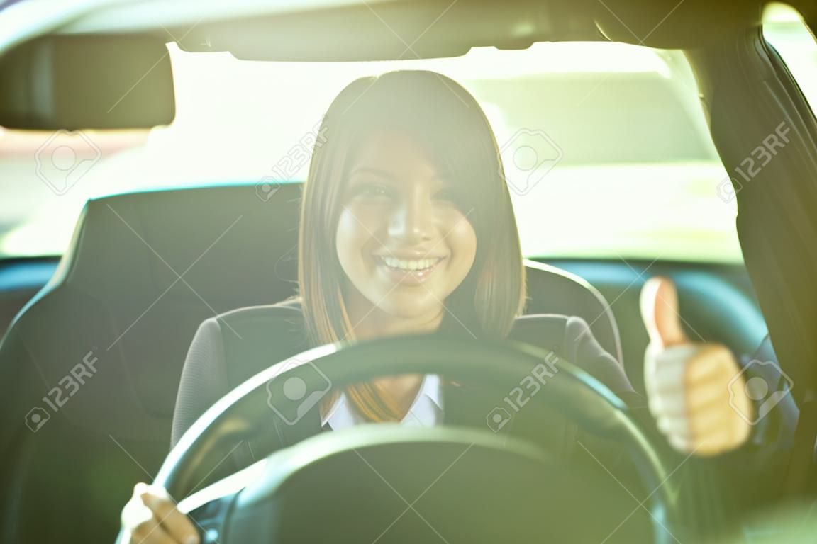 Recht junge Frau, die ein Auto antreibt und lächelnd mit einem Daumen nach oben, durch die Windschutzscheibe gesehen