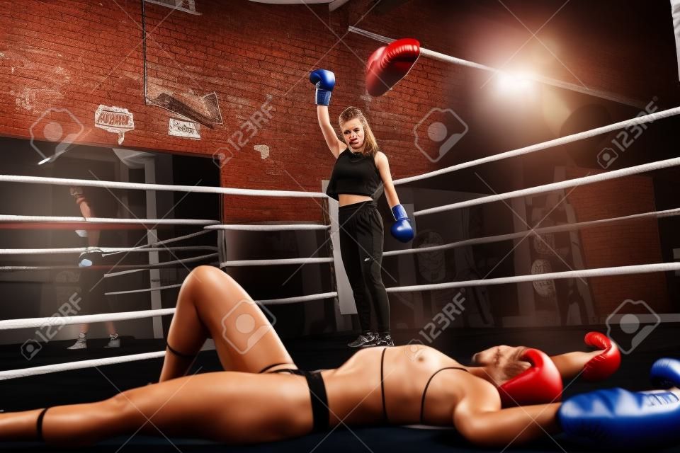 Good looking young woman looking at her defeated opponent after knocking her out on a box fight in a boxing gym