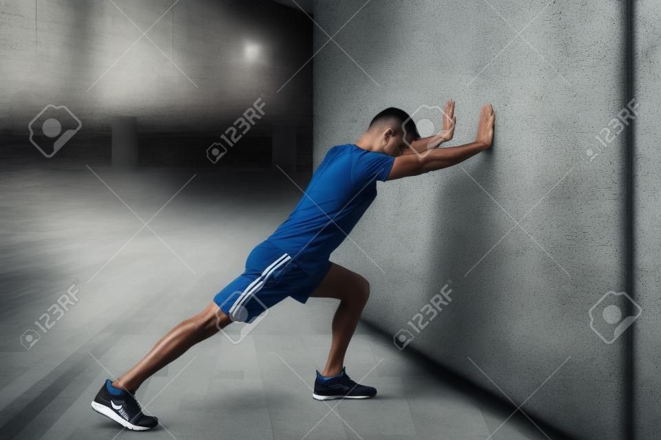 Determined young Latin man stretching calf while leaning on wall