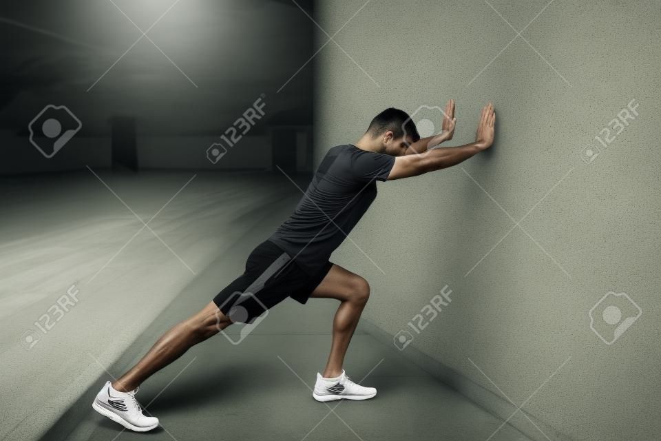 Determined young Latin man stretching calf while leaning on wall