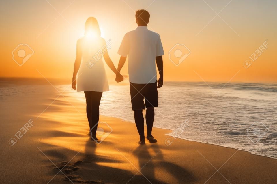 Vue arrière d'un couple marchant main dans la main et sur le sable d'une plage au coucher du soleil