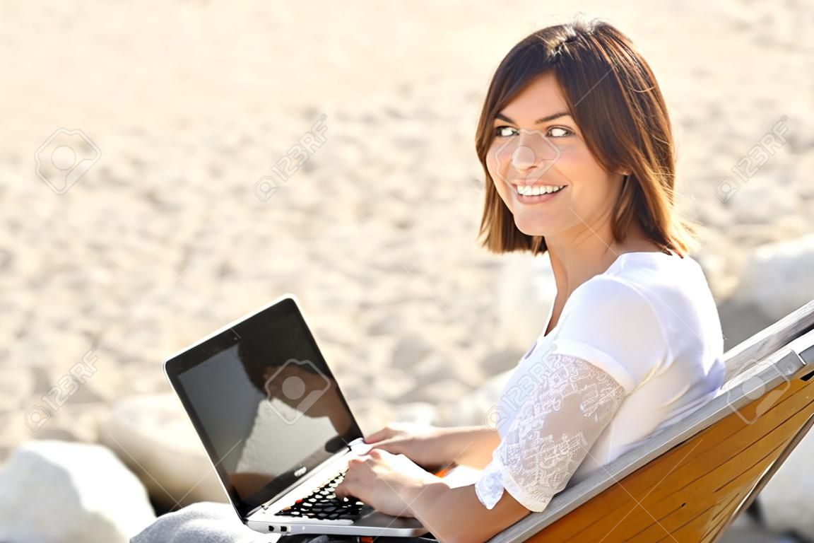Happy woman typing on a laptop and looking at camera sitting on a bench