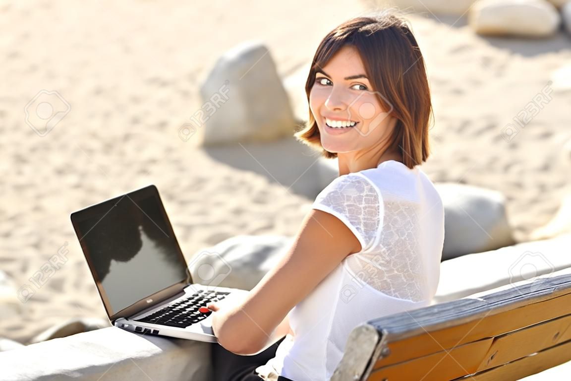 Happy woman typing on a laptop and looking at camera sitting on a bench