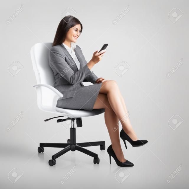 エグゼクティブ ・ ビジネス女性の白い背景に分離された椅子に腰掛けて、スマート フォンを使用して