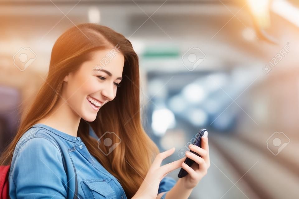 Szczęśliwa dziewczyna tekstylny na inteligentnego telefonu w stacji kolejowej, gdy czeka
