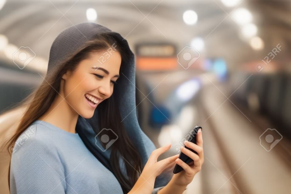 Szczęśliwa dziewczyna tekstylny na inteligentnego telefonu w stacji kolejowej, gdy czeka