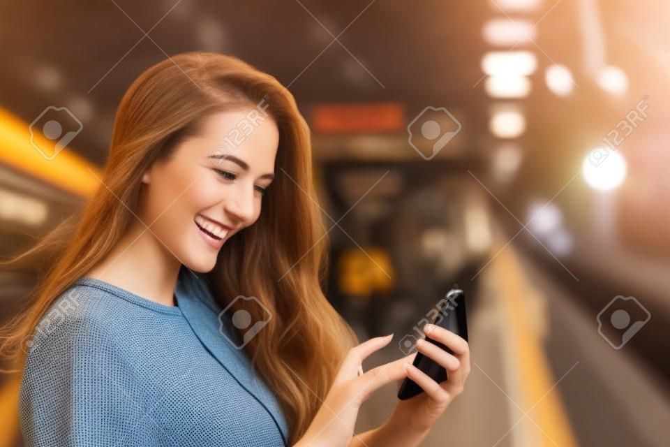 Niña feliz enviando mensajes de texto en un teléfono inteligente en una estación de tren mientras espera