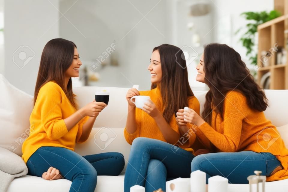 Three happy friends talking and drinking coffee and tea sitting on a couch at home