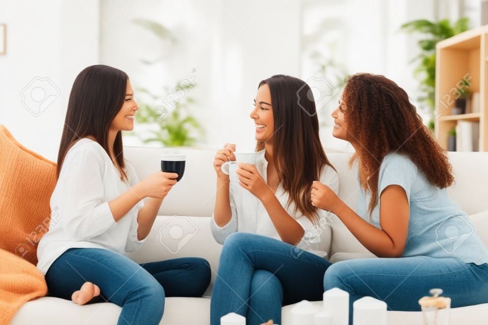 Drei glückliche Freunde reden und trinken Kaffee und Tee sitzt auf einer Couch zu Hause