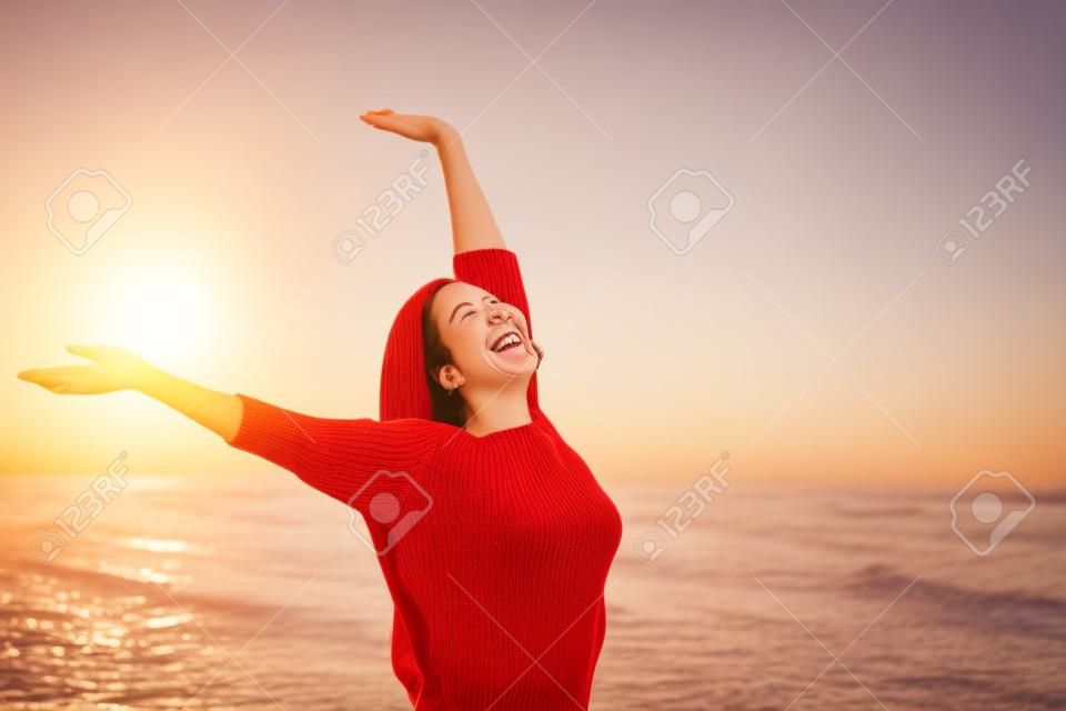 一名快乐的快乐妇女的画象在一个晴朗的早晨激动和举胳膊的穿着在海滩的一件红色毛线衣