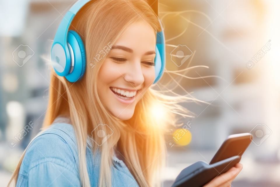 Retrato de una niña feliz escuchando música en línea con auriculares inalámbricos desde un teléfono inteligente en la calle en un día soleado de verano