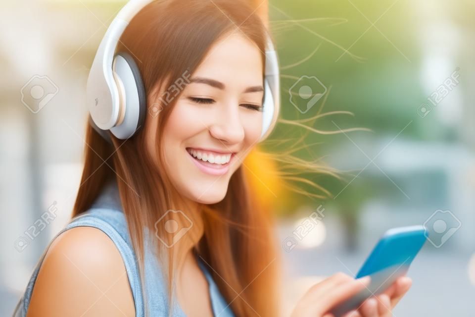 Retrato de una niña feliz escuchando música en línea con auriculares inalámbricos desde un teléfono inteligente en la calle en un día soleado de verano