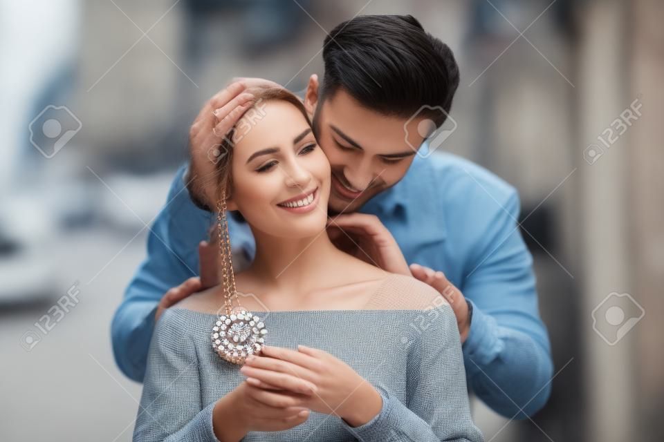 Vista frontal de un hombre que pone una joya collar blanco a su novia en la calle de un lugar romántico. La joyería concepto de modelo.