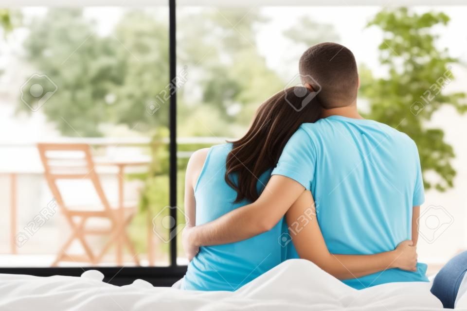 Back view portrait of a happy couple sitting on the bed looking the balcony outdoors through a window of the bedroom of a house