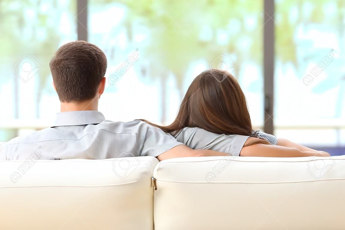 ソファに座って、屋外リビング ルームの窓からの緑の背景を探しているカップルを抱き締めることの後姿